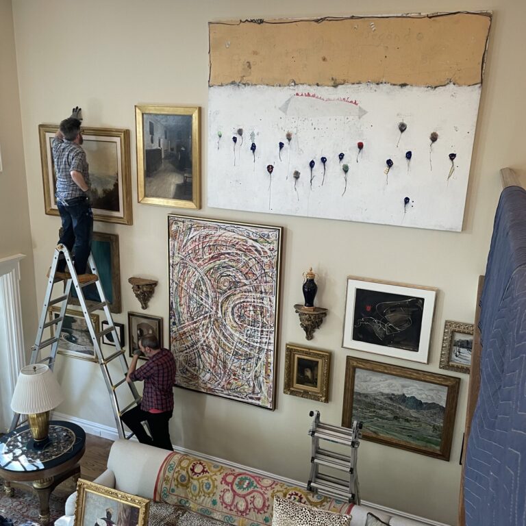 Two art handlers climb a ladder to install and hang large pieces of art onto a yellow wall