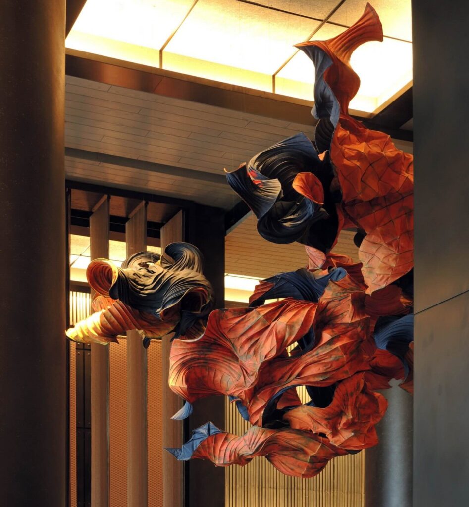 A dramatic large-scale hanging sculpture suspended from a lobby ceiling, featuring sweeping forms in deep rust orange and dark navy that suggest movement and flow, installed in a warmly lit architectural interior.