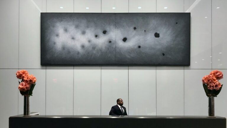 A large horizontal two-panel abstract painting in charcoal and gray tones with dark ovoid forms, professionally installed above a reception desk in a modern corporate lobby.