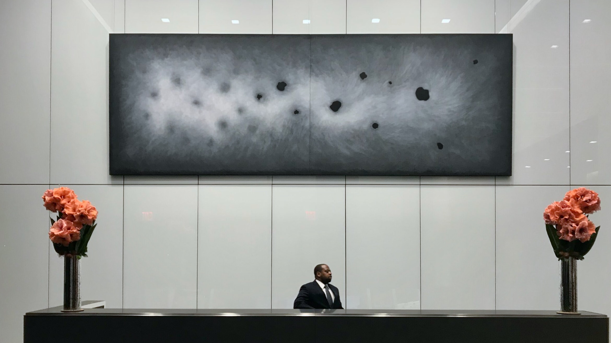 A large horizontal two-panel abstract painting in charcoal and gray tones with dark ovoid forms, professionally installed above a reception desk in a modern corporate lobby.