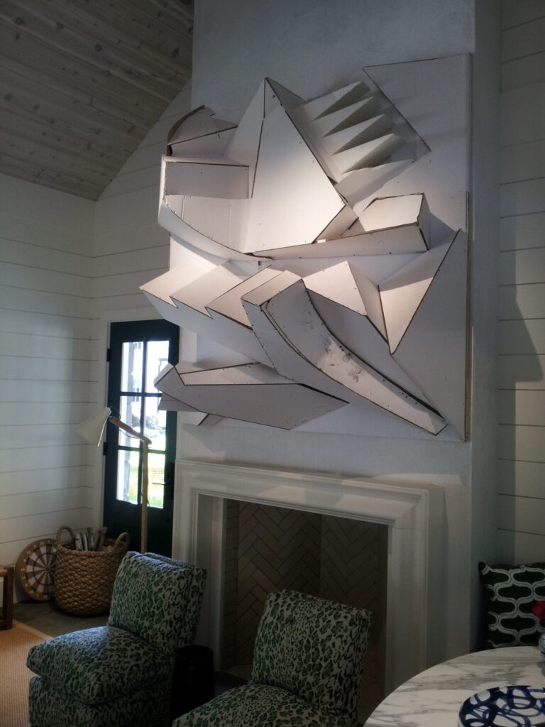 A large abstract three-dimensional white sculptural work with angular, projecting geometric planes, professionally installed on a white shiplap wall above a fireplace in a bright coastal-style living room.