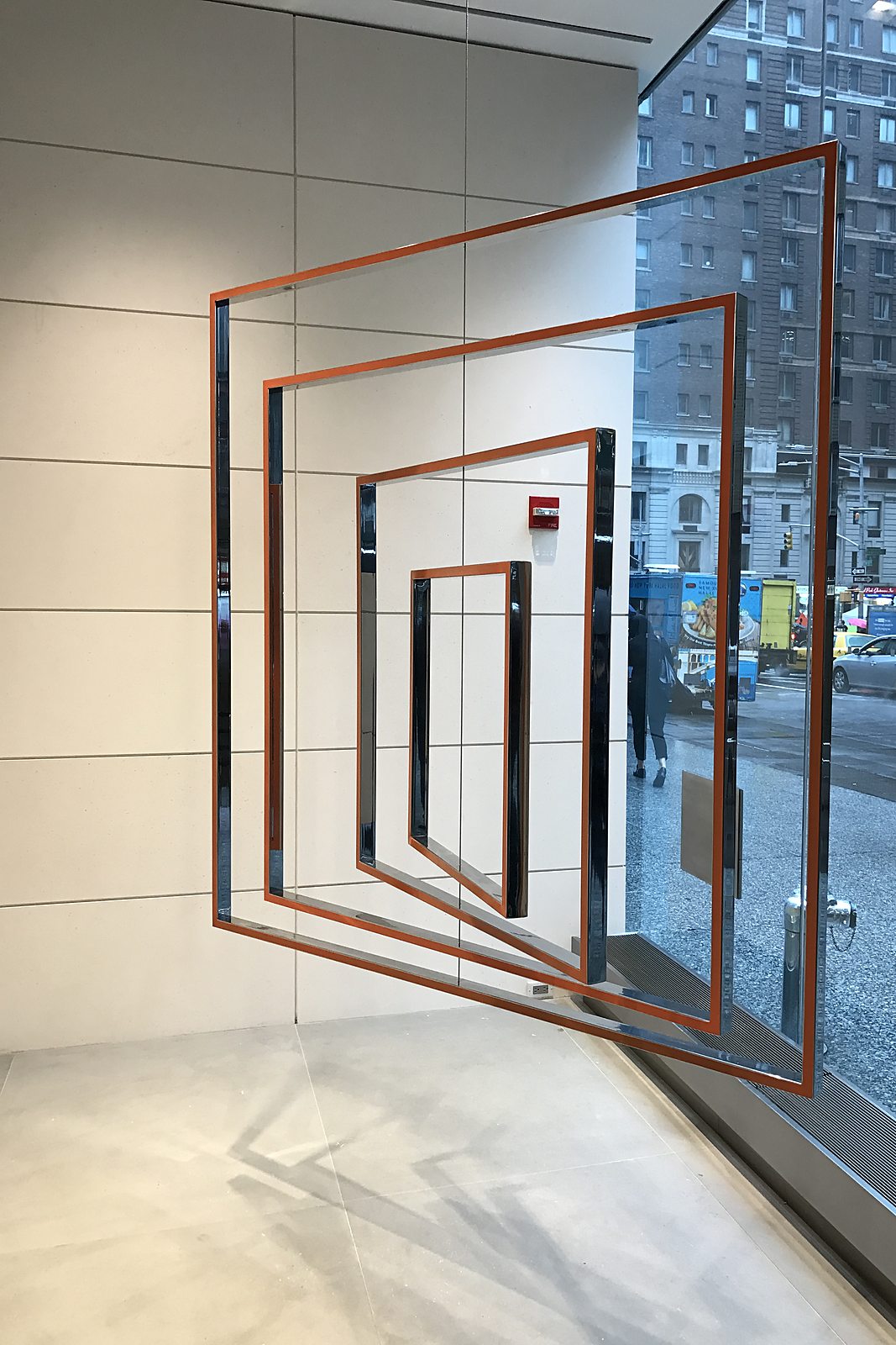 Suspended sculpture of nested orange and steel frames hanging inside a glass building entrance