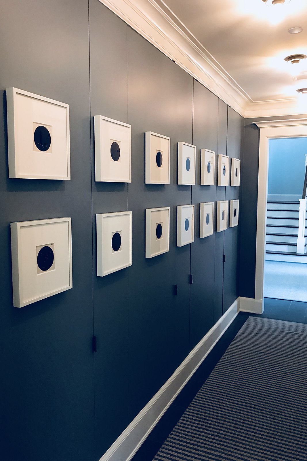 Two rows of white-framed works with dark circular centers installed along a navy corridor