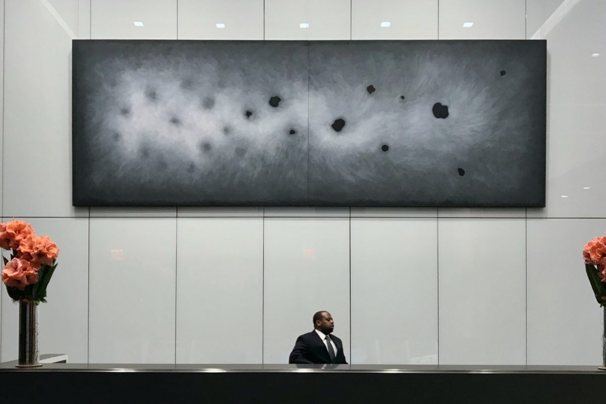 A large horizontal two-panel abstract painting in charcoal and gray tones with dark ovoid forms, professionally installed above a reception desk in a modern corporate lobby.